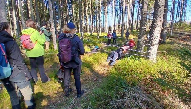 Mežā cilvēki pārgājienā, daži pārvar ap kokiem apsietu striķu šķēršļus, citi skatās